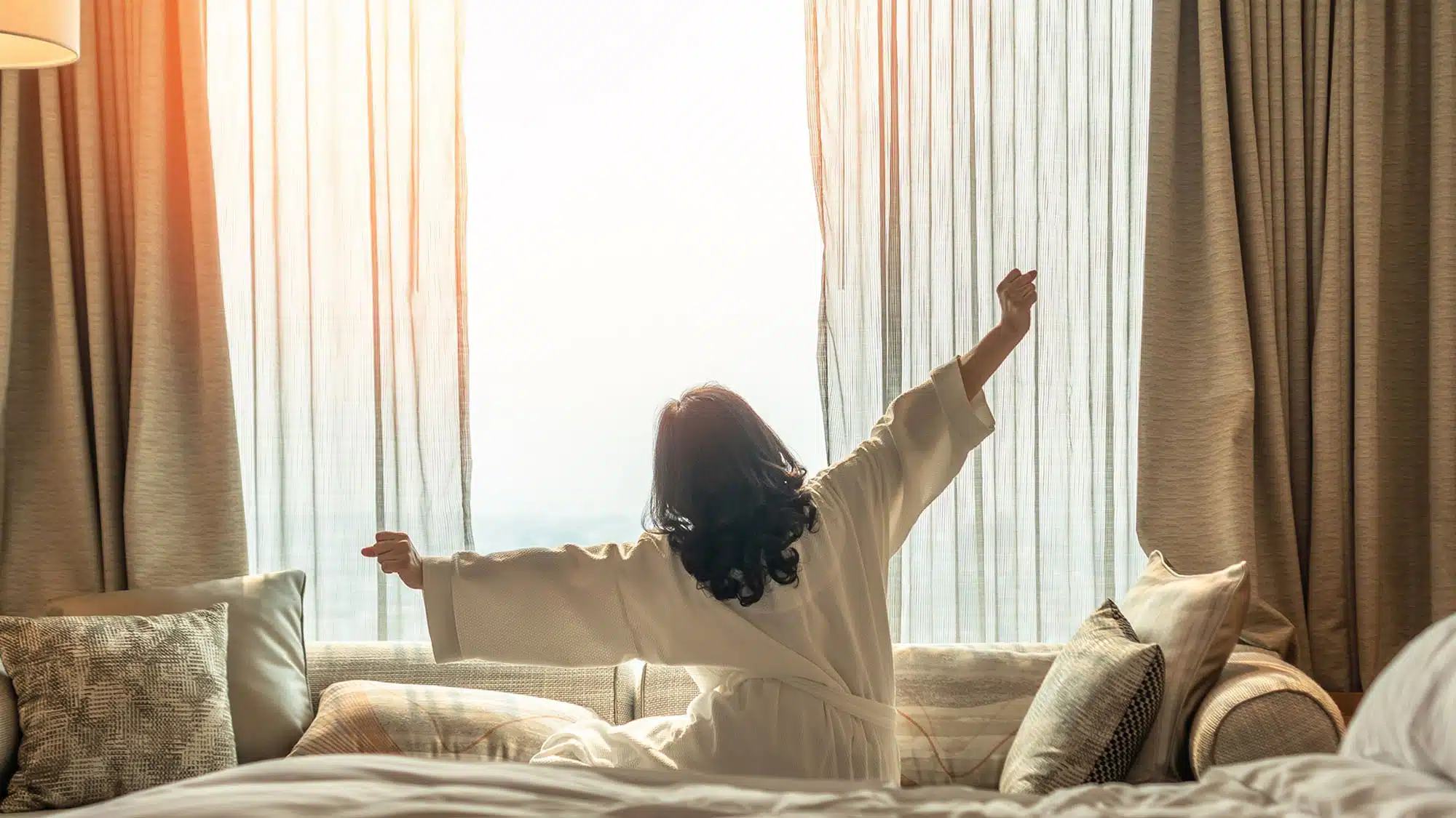 Rückenansicht einer Frau in einem weißen Morgenmantel, die sich nach dem Aufwachen vor einem hellen Fenster mit transparenten Vorhängen streckt. Die sonnendurchflutete Szene symbolisiert die neue Energie und die verbesserte Schlafqualität, die sich bereits wenige Wochen nach dem Rauchstopp mit Asmoken einstellen können.