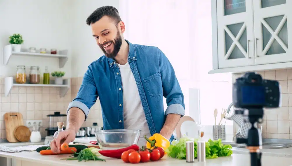Ein lächelnder Mann bereitet in der Küche frisches Gemüse zu. Das Bild zeigt eine gesunde Ernährung als wirksames Mittel gegen Heißhunger während der Nikotinentwöhnung mit Asmoken.