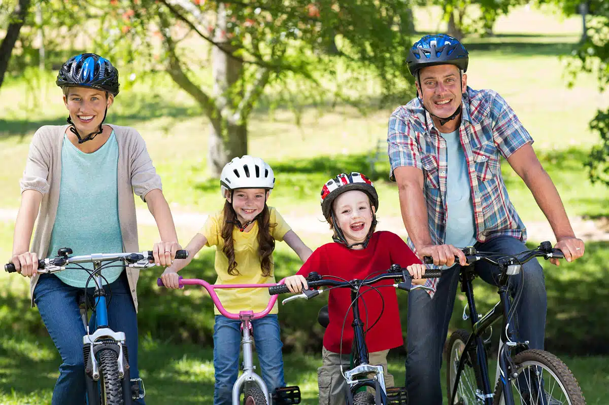 Porträt einer fröhlichen vierköpfigen Familie auf einer Fahrradtour im Park. Alle tragen Helme und lächeln gesundheitsbewusst in die Kamera. Das Bild symbolisiert einen aktiven, rauchfreien Familien-Lifestyle und die wichtige Vorbildfunktion der Eltern für ihre Kinder.