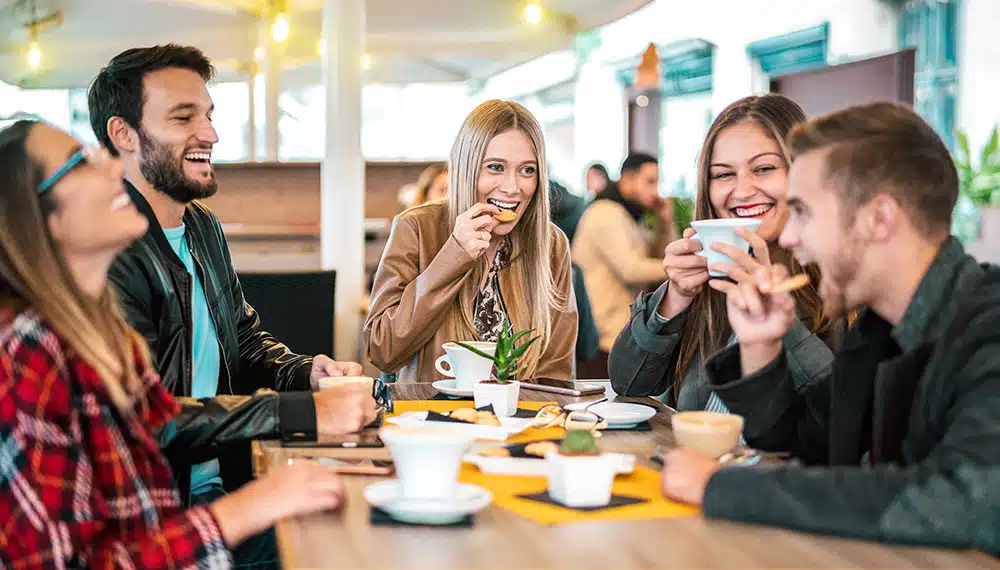 Eine Gruppe von vier jungen Erwachsenen sitzt lachend in einem hellen Café zusammen und unterhält sich angeregt. Das Bild verdeutlicht, wie ein rauchfreies Leben und die Unterstützung durch Freunde die soziale Lebensqualität steigern und andere im Netzwerk zum Rauchstopp mit Asmoken inspirieren können.