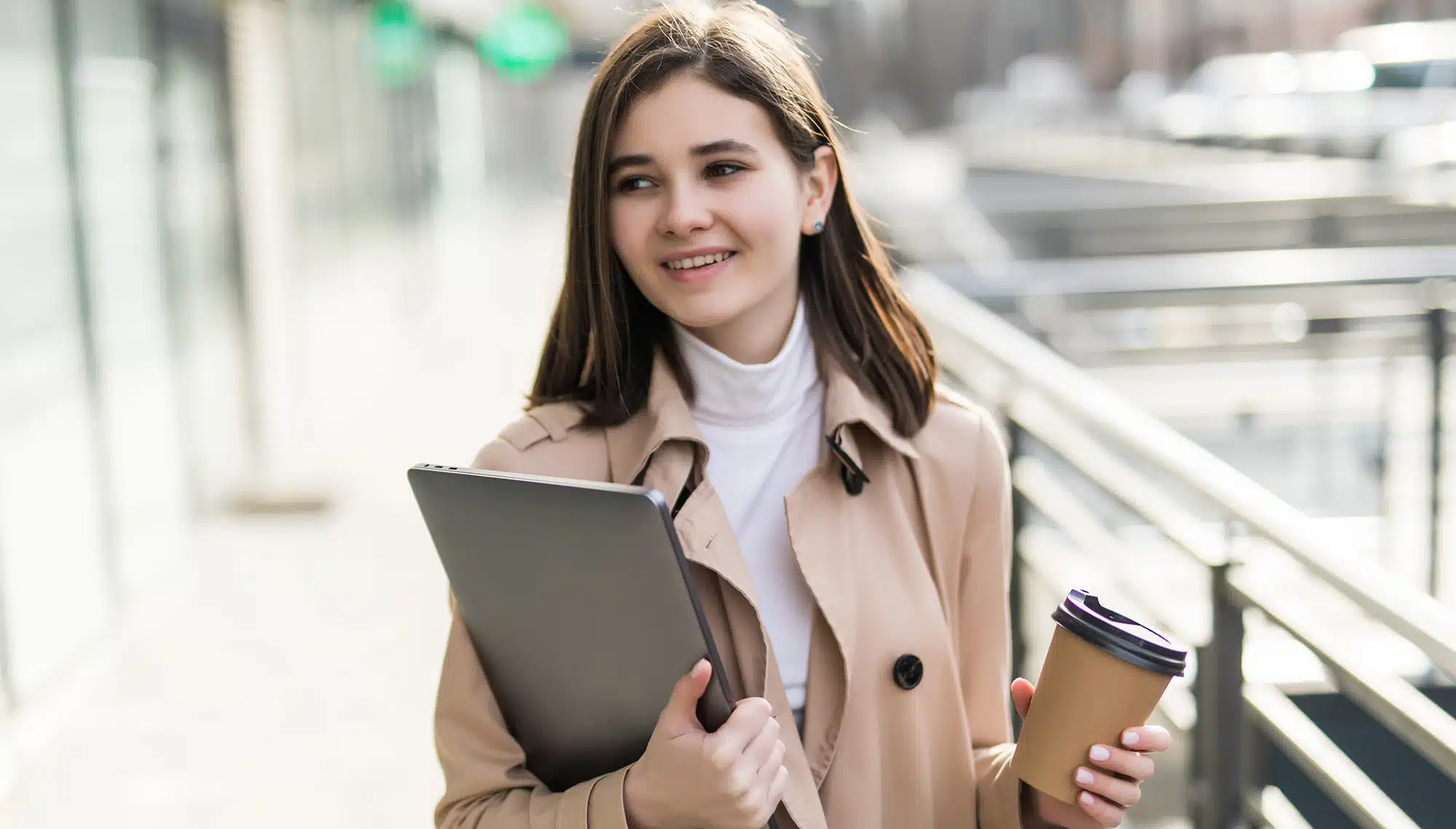 „Nahaufnahme einer lächelnden jungen Frau mit dunklen Haaren, die einen beigen Trenchcoat trägt. Sie hält einen geschlossenen Laptop und einen Coffee-to-go-Becher in den Händen, während sie in einer modernen urbanen Umgebung unterwegs ist. Das Bild symbolisiert einen aktiven, gesunden Lifestyle und den Start in einen rauchfreien neuen Lebensabschnitt.
