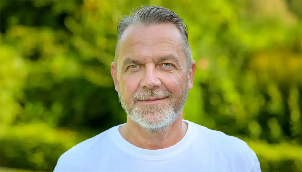 Nahaufnahme von G&uuml;nter mit kurzem grauen Haar und gepflegtem Dreitagebart, bekleidet mit einem wei&szlig;en T-Shirt vor gr&uuml;nem Hintergrund. Als ehemaliger Raucher mit &uuml;ber 30 Jahren Konsumerfahrung teilt er seine Geschichte &uuml;ber gesundheitliche Beschwerden und den endg&uuml;ltigen Rauchstopp mit Asmoken.