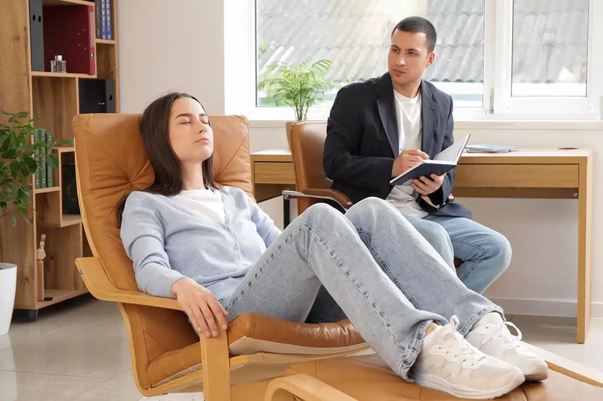 „Eine junge Frau liegt entspannt mit geschlossenen Augen auf einem hellbraunen Loungesessel. Neben ihr sitzt ein Therapeut und macht sich Notizen. Das Bild symbolisiert eine Sitzung zur mentalen Unterstützung während der Raucherentwöhnung, die durch Hypnose oder medikamentöse Ansätze wie Asmoken ergänzt werden kann.