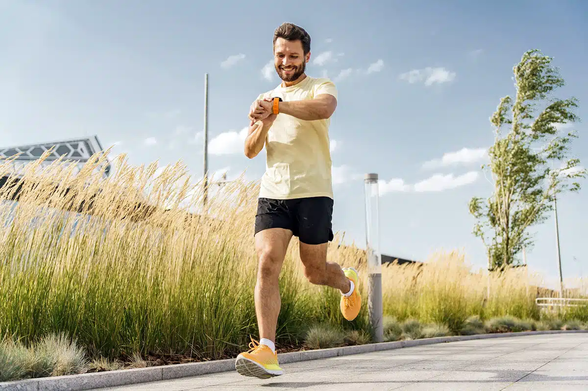 „Ein Mann beim Joggen auf einem modernen urbanen Weg unter klarem Himmel. Er blickt lächelnd auf seine Fitness-Smartwatch am Handgelenk. Das Bild visualisiert die wiedergewonnene Leistungsfähigkeit und die positiven gesundheitlichen Fortschritte, die durch einen Rauchstopp mit Unterstützung von Asmoken möglich werden.