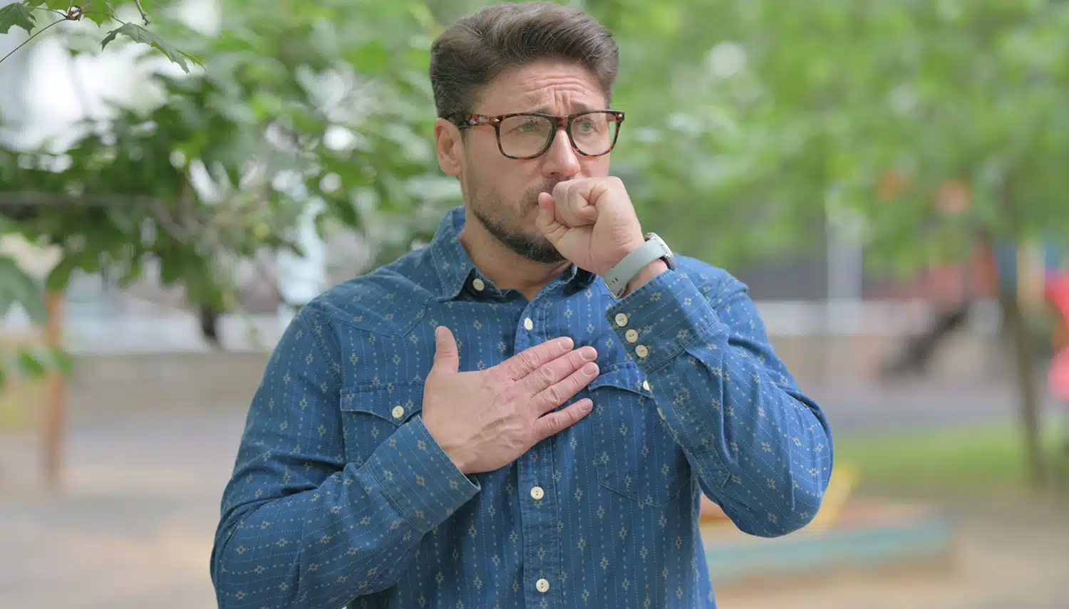 Porträt eines Mannes mittleren Alters mit Bart und Brille, der in einem Park steht, hustet und sich die Hand an die Brust hält. Das Bild symbolisiert körperliche Warnsignale durch Rauchen wie Husten und Lungenbeschwerden, die oft der Auslöser für eine Raucherentwöhnung mit Asmoken sind.