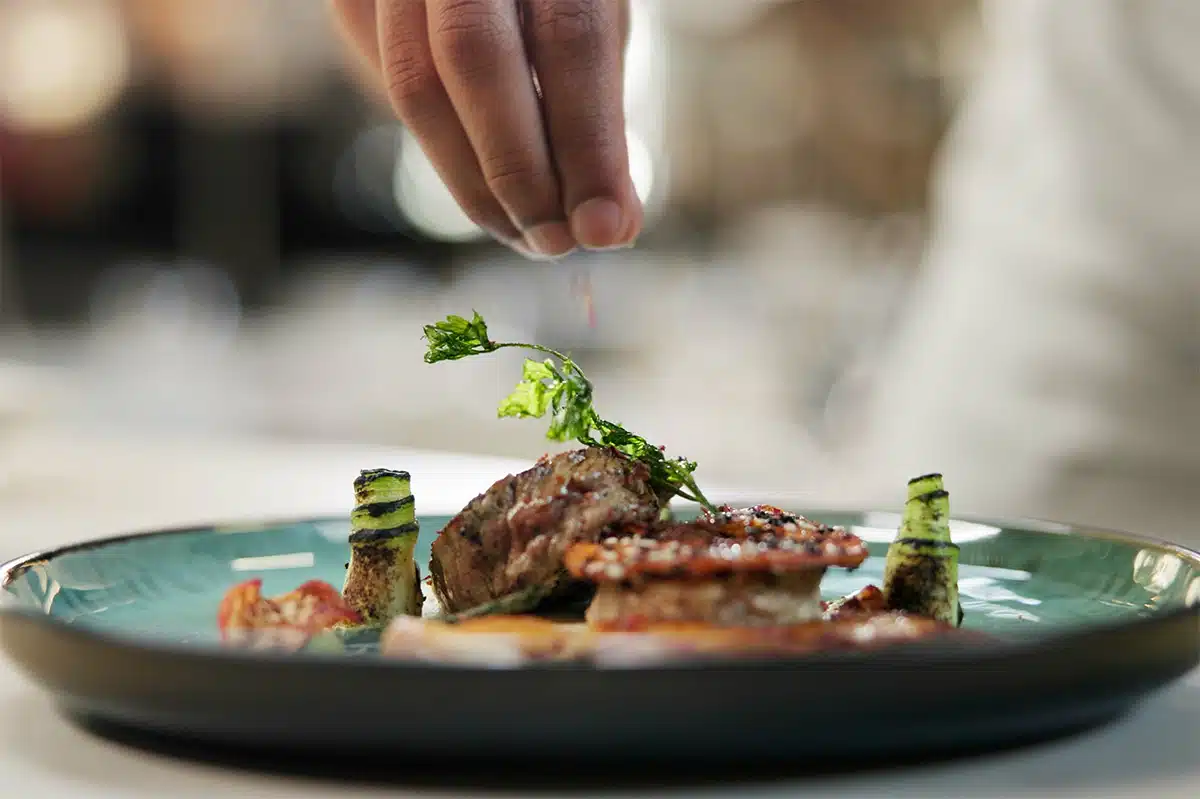 Nahaufnahme einer Hand, die eine Prise Gewürz über ein kunstvoll angerichtetes Gourmet-Gericht mit Fleisch und Zucchini-Röllchen streut. Das Bild symbolisiert die Wiederentdeckung des Geschmackssinns und den Fokus auf gesunde Ernährung nach einem erfolgreichen Rauchstopp mit Asmoken.