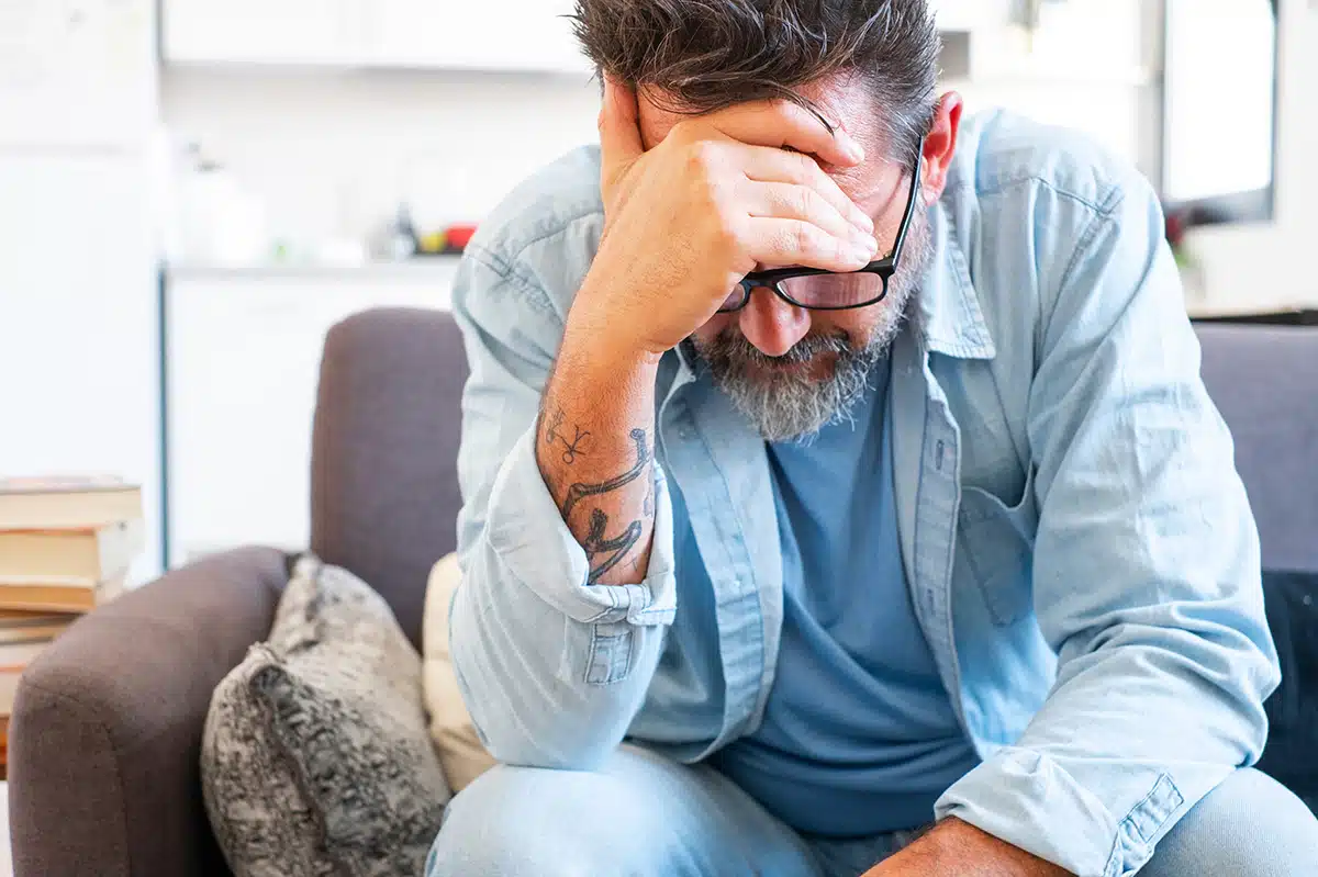 Nahaufnahme eines Mannes mit Brille und Bart, der verzweifelt den Kopf in die Hände stützt. Das Bild symbolisiert die psychische Belastung, Stress und das Verlangen während des Nikotinentzugs, dem man mit der Asmoken-Kur in 25 Tagen entgegenwirken kann.