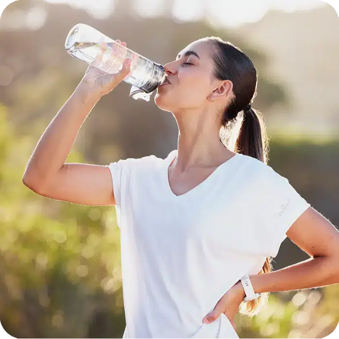 Eine junge Frau mit zum Pferdeschwanz gebundenen Haaren trinkt in einer sonnigen Außenumgebung Wasser aus einer klaren Sportflasche. Das Bild symbolisiert gesunde Gewohnheiten und die wichtige Flüssigkeitszufuhr zur Unterstützung der Entgiftung während der Raucherentwöhnung mit Asmoken.