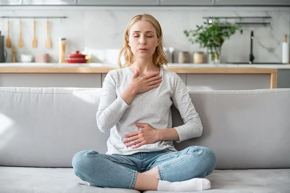 Eine junge Frau sitzt im Schneidersitz mit geschlossenen Augen auf einem Sofa und praktiziert eine Beruhigungsübung, indem sie eine Hand auf ihre Brust und die andere auf ihren Bauch legt. Dies illustriert Entspannungstechniken zur Bewältigung von Stressmomenten während der Raucherentwöhnung.