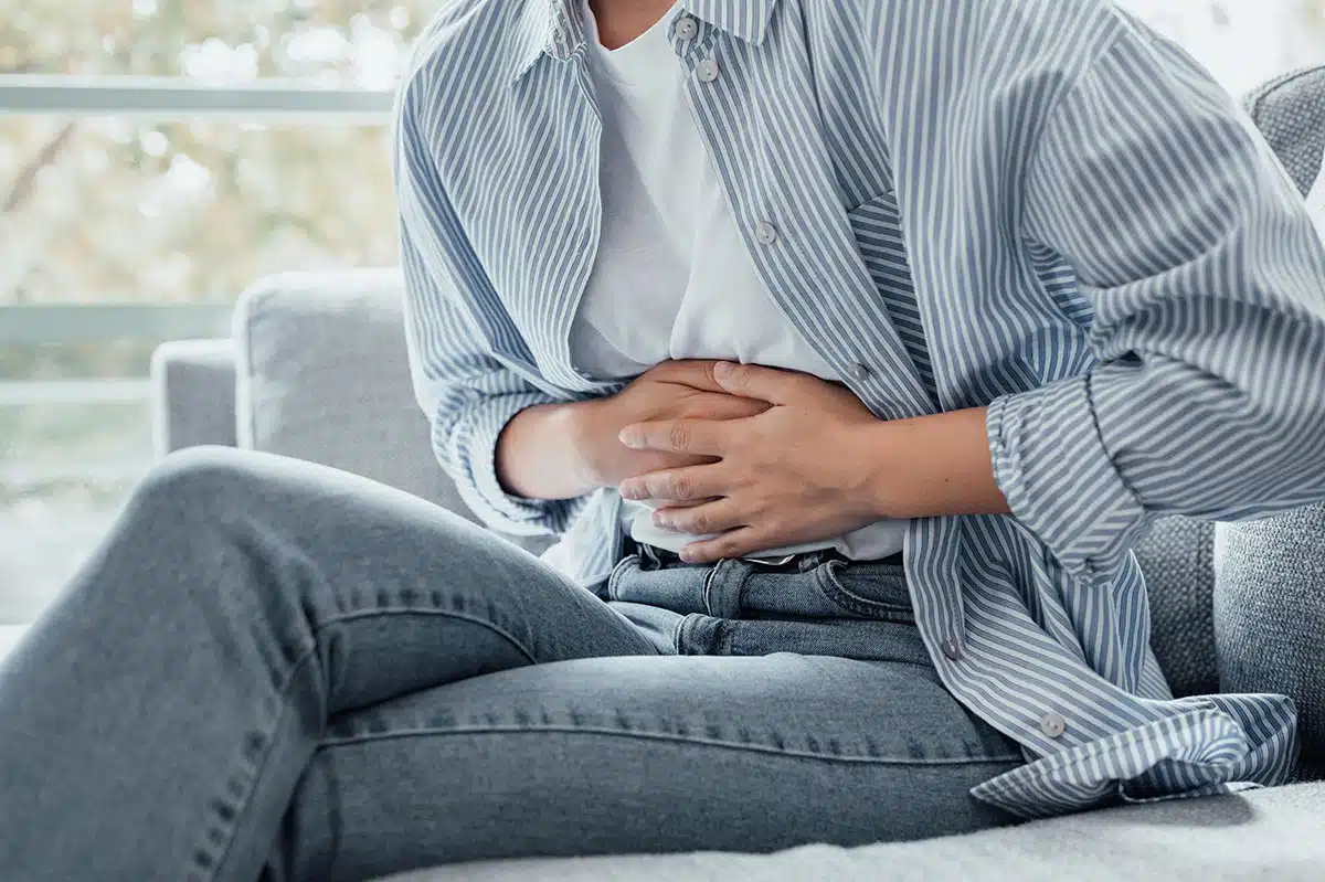 ahaufnahme einer Person in einem gestreiften Hemd, die auf einem Sofa sitzt und sich mit beiden Händen den Bauch hält. Das Bild verdeutlicht den Zusammenhang zwischen Rauchen und Verdauungsbeschwerden sowie die Bedeutung der Darmgesundheit, die durch einen Rauchstopp mit Asmoken wiederhergestellt werden kann.