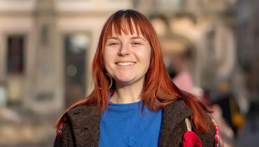 Porträt der 25-jährigen Anna mit langen roten Haaren und Pony, die in einem blauen Oberteil und dunkler Jacke freundlich lächelt. Als Pharmazeutisch-kaufmännische Angestellte (PKA) teilt sie ihre fachliche und persönliche Erfahrung mit Asmoken.