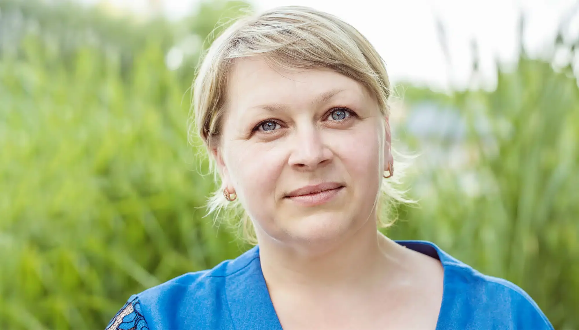„Nahaufnahme von Sandra (42), einer Frau mit blonden, kurz geschnittenen Haaren und blauen Augen, die ein blaues Oberteil trägt. Vor einem natürlichen, grünen Hintergrund blickt sie ruhig in die Kamera. Als ehemalige Raucherin teilt sie ihre Geschichte über die jahrzehntelange Abhängigkeit und ihren erfolgreichen Weg mit Asmoken.